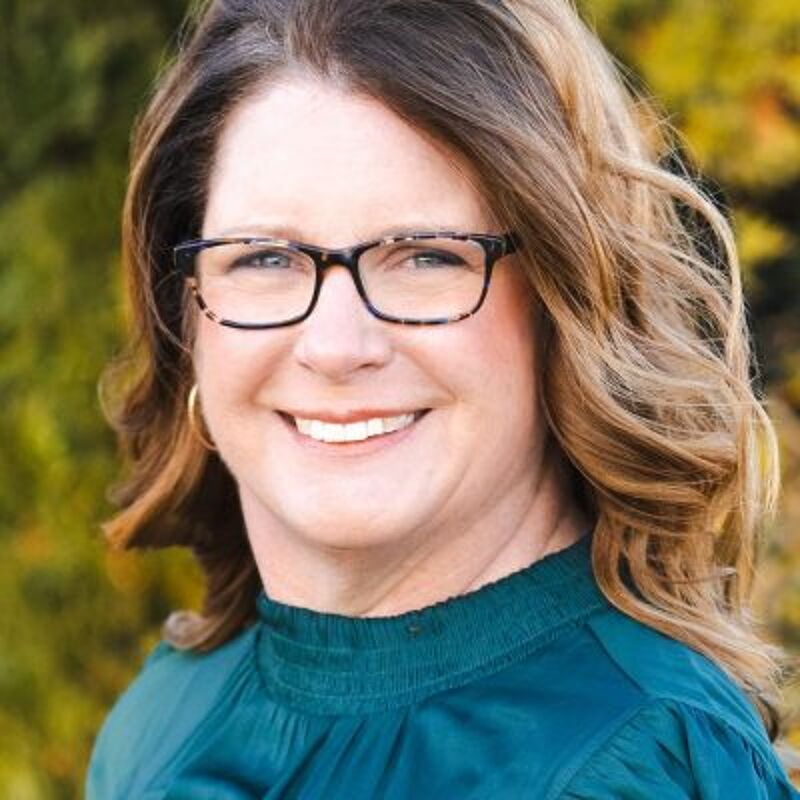The image shows a woman with glasses smiling at the camera. She has brown hair and is wearing a green shirt. The background is blurred, suggesting an outdoor setting. She appears to be in her 40s or 50s. The overall impression is friendly and approachable.